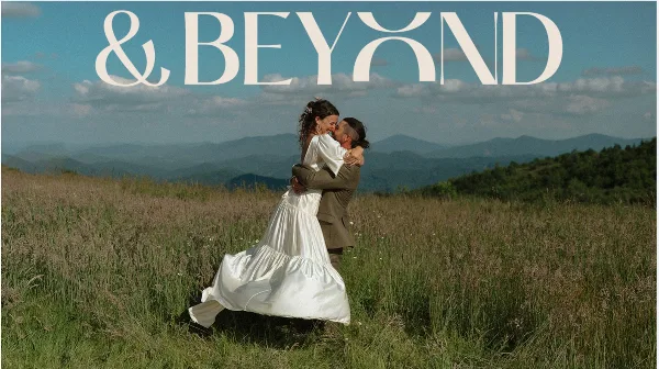 A bride and groom embracing in a grassy field with mountains in the background, with the text '& BEYOND' overlaid at the top.
