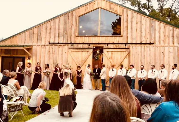 A wedding ceremony in progress with a bride and groom at the altar, surrounded by wedding party members, in front of a large wooden barn.