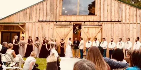 A wedding ceremony in progress with a bride and groom at the altar, surrounded by wedding party members, in front of a large wooden barn.