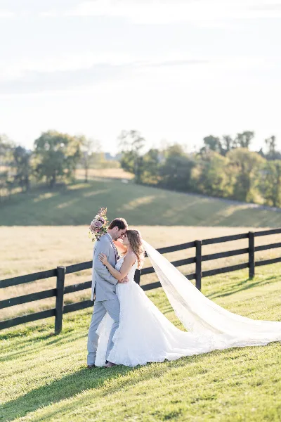 A bride and groom embracing in a grassy field with a wooden fence and trees in the background.