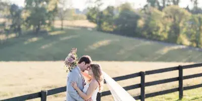 A bride and groom embracing in a grassy field with a wooden fence and trees in the background.