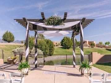 A wooden pergola decorated with white draping and floral arrangements at an outdoor event venue.