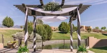 A wooden pergola decorated with white draping and floral arrangements at an outdoor event venue.