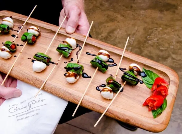 A person holding a wooden serving board with skewered mozzarella, basil, and tomato appetizers drizzled with balsamic glaze, next to a Catering By Design branded napkin.