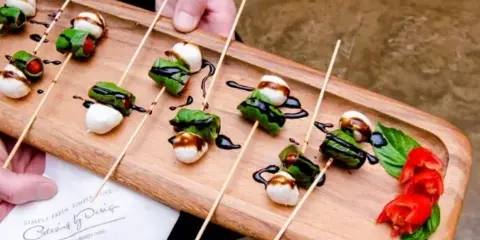 A person holding a wooden serving board with skewered mozzarella, basil, and tomato appetizers drizzled with balsamic glaze, next to a Catering By Design branded napkin.