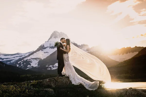 A bride and groom kissing on a rocky outcrop with a snow-capped mountain and lake in the background at sunset.