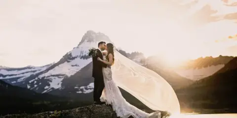 A bride and groom kissing on a rocky outcrop with a snow-capped mountain and lake in the background at sunset.