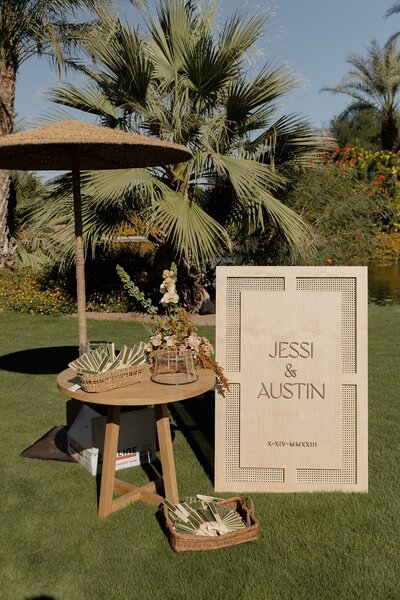 A wooden wedding welcome sign for 'Jessi & Austin' stands on a lawn next to a small table with floral arrangements and palm fronds, set against a backdrop of palm trees.