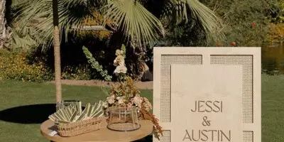 A wooden wedding welcome sign for 'Jessi & Austin' stands on a lawn next to a small table with floral arrangements and palm fronds, set against a backdrop of palm trees.
