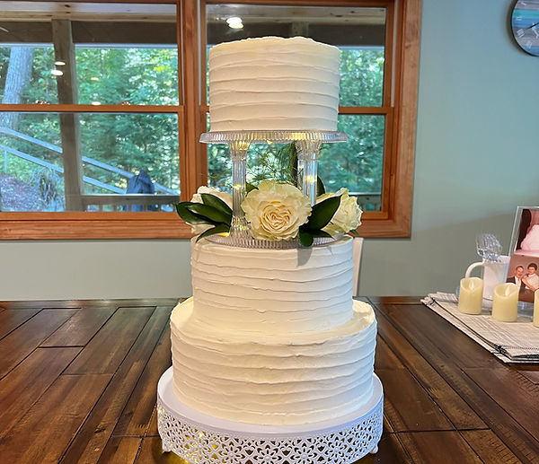 Three-tier white buttercream wedding cake with white roses and greenery on a clear acrylic stand, displayed on a wooden table.