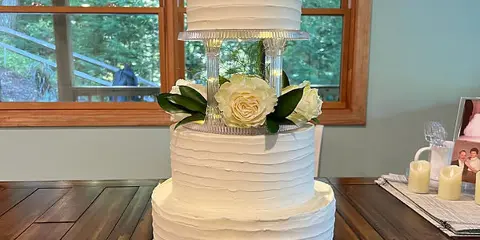 Three-tier white buttercream wedding cake with white roses and greenery on a clear acrylic stand, displayed on a wooden table.