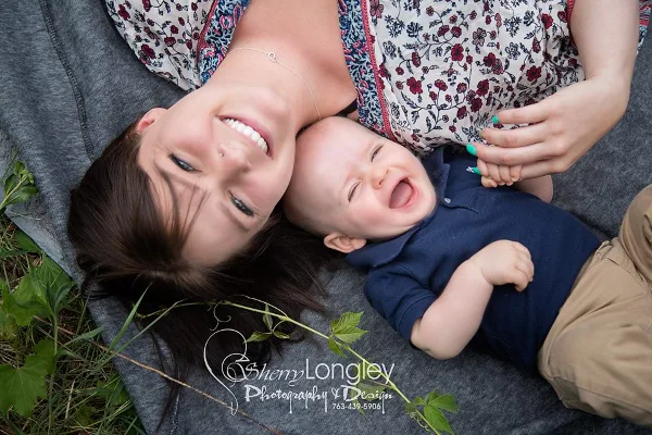 A smiling mother and a laughing baby lying on a grey blanket outdoors, with a Sherry Longley Photography watermark.