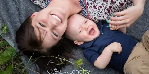 A smiling mother and a laughing baby lying on a grey blanket outdoors, with a Sherry Longley Photography watermark.