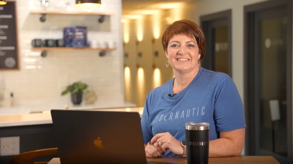 A woman in a blue t-shirt sitting at a desk with a laptop, filmed in a professional office or cafe setting.