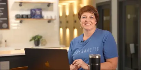 A woman in a blue t-shirt sitting at a desk with a laptop, filmed in a professional office or cafe setting.