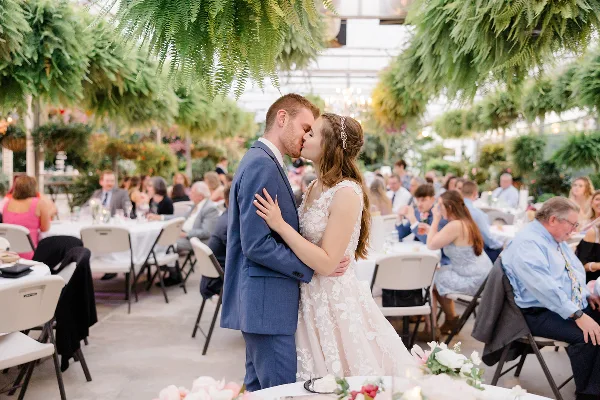 A bride and groom sharing a kiss during a wedding reception inside a greenhouse conservatory filled with hanging ferns and lush greenery.