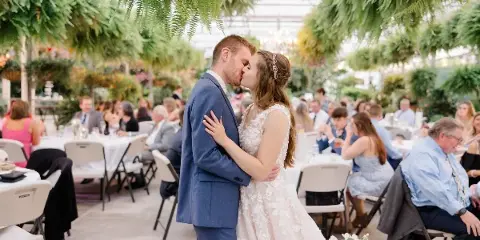 A bride and groom sharing a kiss during a wedding reception inside a greenhouse conservatory filled with hanging ferns and lush greenery.