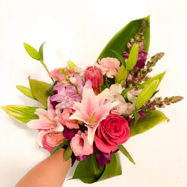 A hand holding a mixed floral bouquet featuring pink lilies, roses, and orchids against a white background.