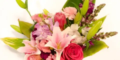A hand holding a mixed floral bouquet featuring pink lilies, roses, and orchids against a white background.