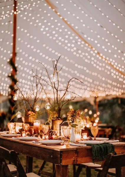 A professionally styled wedding reception table featuring floral centerpieces, candles, and string lights under a tent.