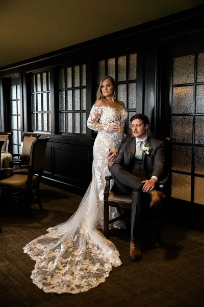 A bride in a lace wedding gown and a groom in a suit posing in a wood-paneled room.