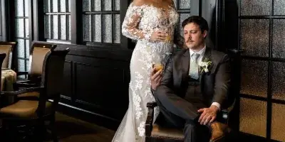 A bride in a lace wedding gown and a groom in a suit posing in a wood-paneled room.