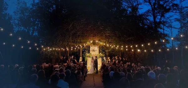 Outdoor wedding ceremony at dusk with a bride and groom at a decorated altar under string lights, surrounded by guests.