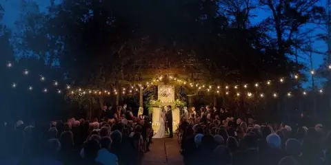 Outdoor wedding ceremony at dusk with a bride and groom at a decorated altar under string lights, surrounded by guests.
