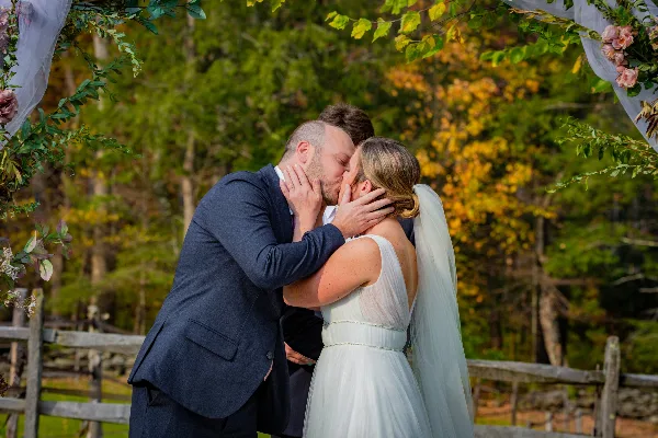 A bride and groom kissing at an outdoor wedding ceremony with a floral arch and autumn trees in the background.