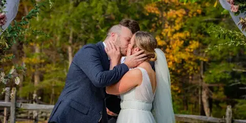 A bride and groom kissing at an outdoor wedding ceremony with a floral arch and autumn trees in the background.