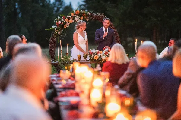A bride and groom standing at a floral arch during an outdoor wedding ceremony with a candlelit table in the foreground.