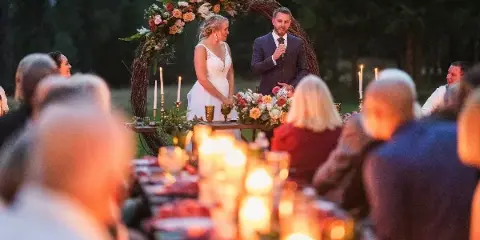 A bride and groom standing at a floral arch during an outdoor wedding ceremony with a candlelit table in the foreground.