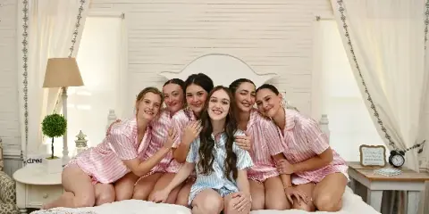 A group of women in matching pink and blue striped pajamas posing on a bed in a white-walled guest room with a 'Welcome to our home' sign.