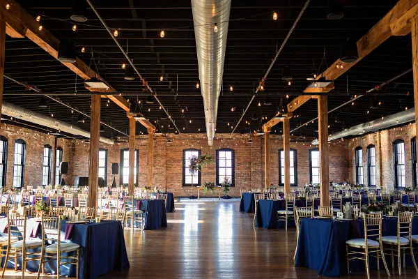 A spacious industrial-style wedding venue featuring exposed brick walls, wooden beams, and round tables set with blue linens and floral centerpieces.