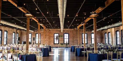A spacious industrial-style wedding venue featuring exposed brick walls, wooden beams, and round tables set with blue linens and floral centerpieces.
