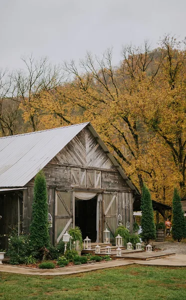 Exterior view of a rustic wooden barn wedding venue with a metal roof, surrounded by evergreen trees and autumn foliage.