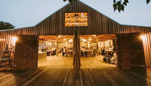 Exterior view of a wooden barn venue with open doors showing a reception area with guests and tables.