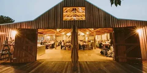 Exterior view of a wooden barn venue with open doors showing a reception area with guests and tables.