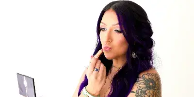 A woman with purple hair and floral tattoos applying lipstick while looking into a compact mirror.