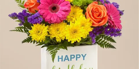 Colorful floral arrangement with pink gerberas, yellow daisies, orange roses, and purple stock in a white box labeled 'Happy Birthday' with a red ribbon.