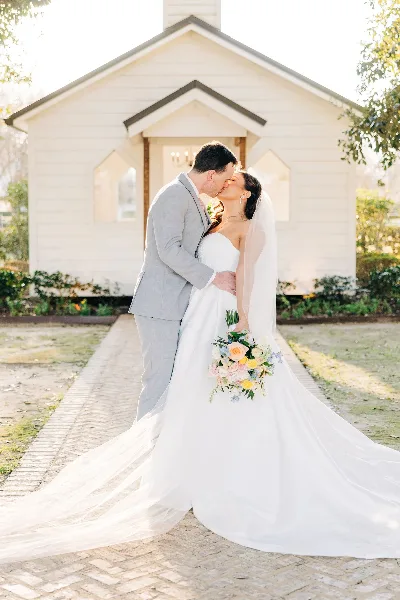 A bride and groom kissing in front of a white chapel building, with the bride holding a colorful bouquet.