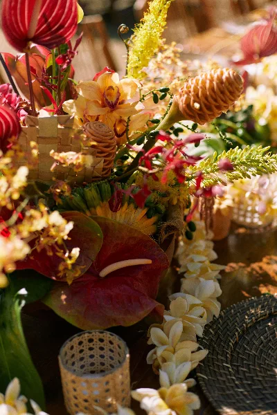 A close-up of a tropical floral arrangement featuring red anthuriums, yellow orchids, and a white plumeria lei resting on a woven mat.
