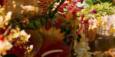 A close-up of a tropical floral arrangement featuring red anthuriums, yellow orchids, and a white plumeria lei resting on a woven mat.