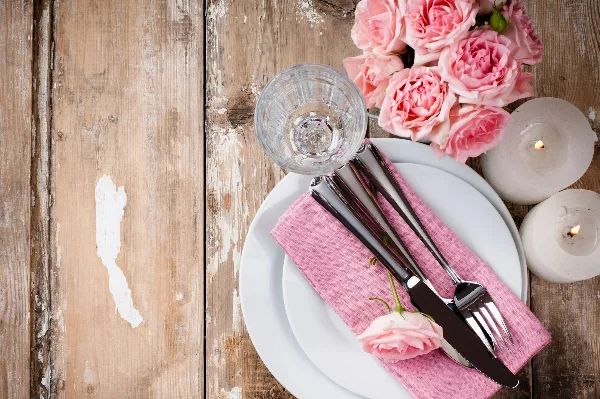 A rustic wedding table setting featuring a white plate, pink napkin, silverware, a glass, pink roses, and lit candles on a distressed wooden surface.