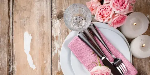 A rustic wedding table setting featuring a white plate, pink napkin, silverware, a glass, pink roses, and lit candles on a distressed wooden surface.