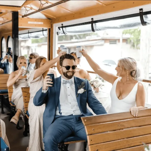 A bride and groom in formal wedding attire sitting on a wooden trolley with guests, raising drinks in a toast.