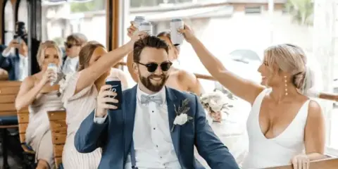 A bride and groom in formal wedding attire sitting on a wooden trolley with guests, raising drinks in a toast.