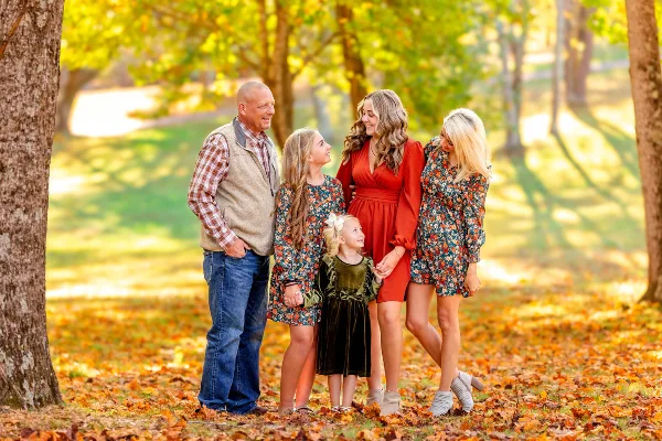 A professional family portrait of a man, two women, and two young girls standing together in a park with autumn leaves.