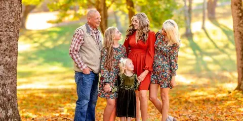 A professional family portrait of a man, two women, and two young girls standing together in a park with autumn leaves.