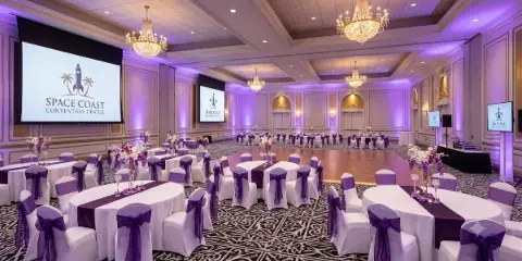 A large indoor ballroom set up for a wedding reception with round tables, purple chair sashes, floral centerpieces, and a wooden dance floor.